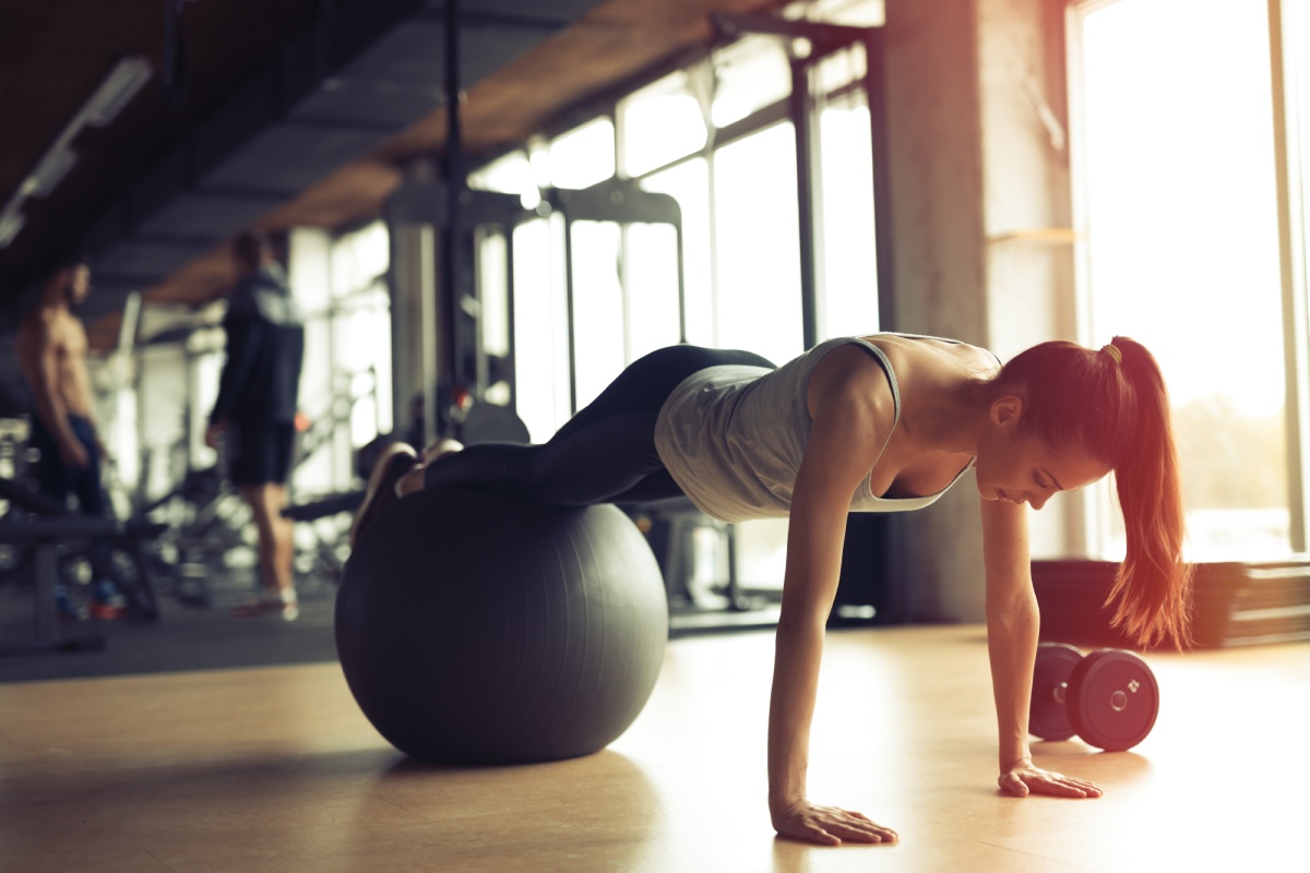 A person exercises in a gym by balancing their legs on a stability ball while doing a plank. A dumbbell rests nearby. Other gym-goers and equipment are visible in the background.