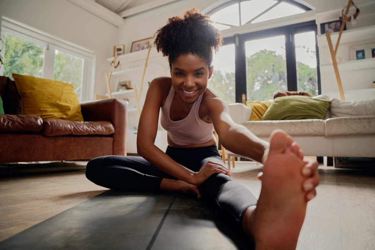 A person sitting on a yoga mat indoors, stretching with one leg extended and holding their foot, smiling at the camera. A sofa with colorful cushions is visible in the background.