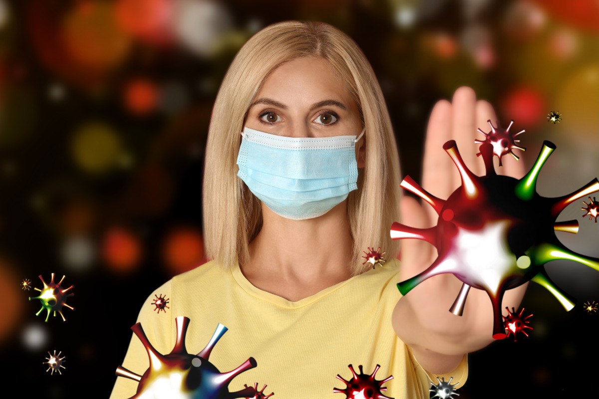 A woman wearing a face mask holds up her hand in a stop gesture as illustrated viruses surround her.