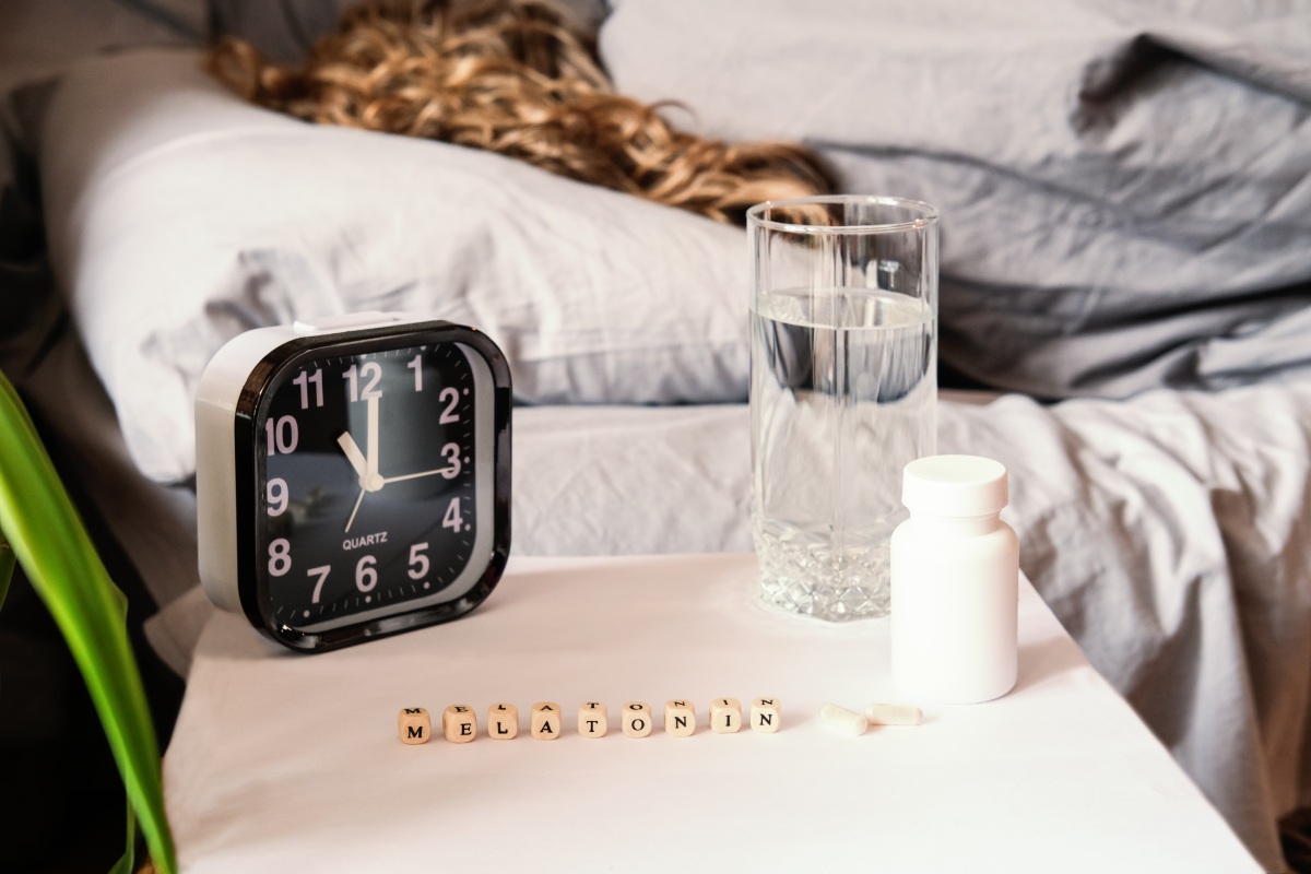 A bedside table with an alarm clock showing 4:15, a glass of water, a bottle of pills, scattered pills, and blocks spelling "melatonin." A person with blonde hair is sleeping in bed in the background.