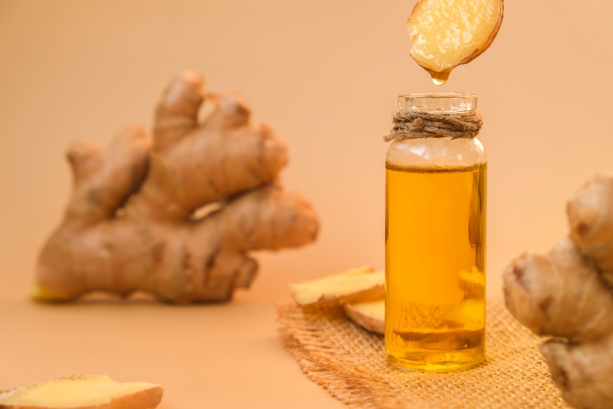A bottle filled with golden liquid, surrounded by ginger roots and ginger slices. One ginger slice above the bottle drips liquid into it. Set against a beige background.