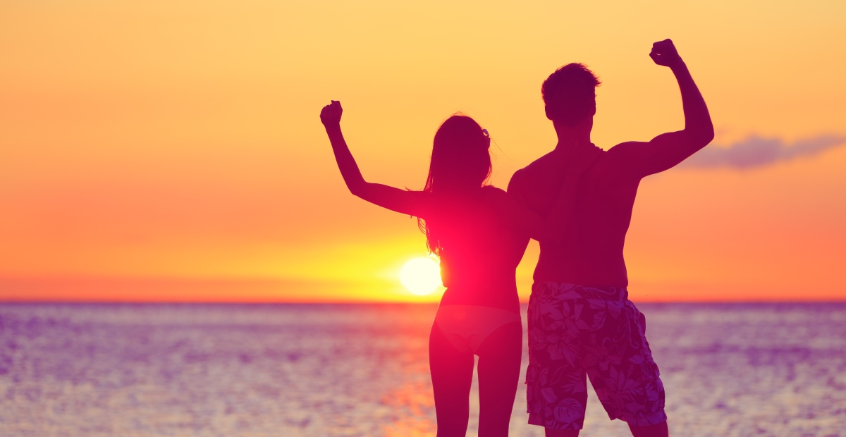Silhouetted couple in swimwear stands with arms raised against a vibrant orange and pink sunset over the ocean.