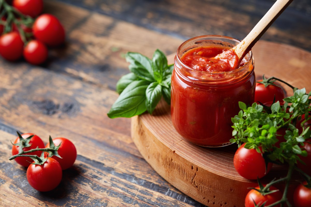 A jar of tomato sauce with a wooden spoon sits on a wooden surface. Fresh basil and clusters of tomatoes surround the jar.