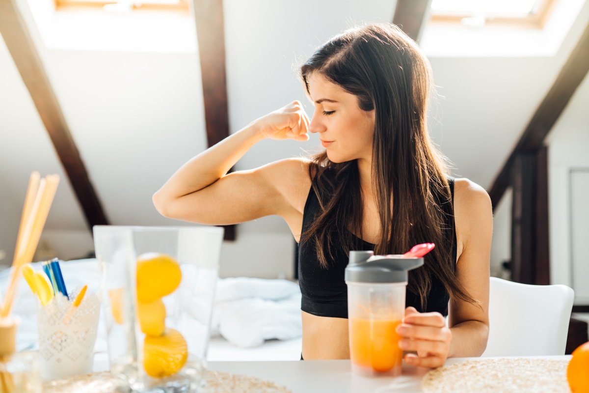 A woman in a black sports bra sits at a table, flexing her bicep with her left arm while holding a shaker bottle with an orange drink in her right hand. Lemons and kitchen items are on the table.