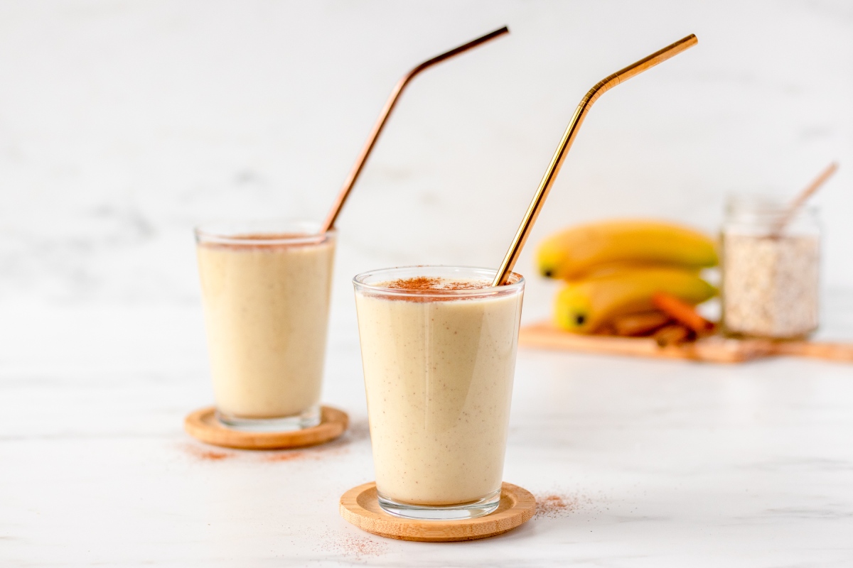 Two glasses of banana smoothies with metal straws on wooden coasters. In the background, there are bananas, a jar of oats, and cinnamon sticks on a wooden tray.