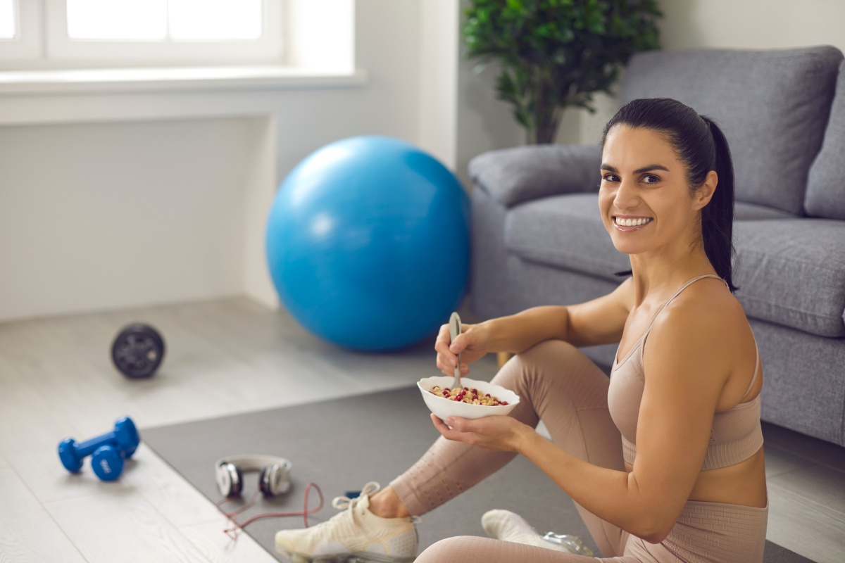 A woman in workout attire sits on a mat indoors, smiling and holding a bowl of food. Fitness equipment, including a blue exercise ball and dumbbells, is visible nearby.