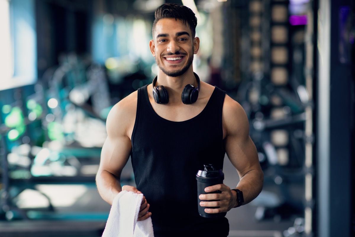 Man at the gym in a black tank top holding a water bottle and a towel, wearing headphones around his neck, and smiling. Fitness equipment is visible in the background.