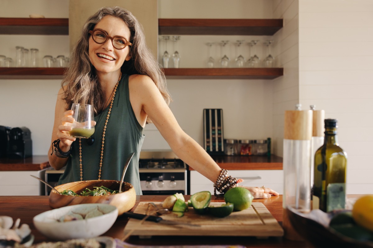 A smiling woman with long gray hair and glasses stands in a kitchen, holding a drink. A cutting board with avocado, lime, and vegetables is on the counter in front of her.
