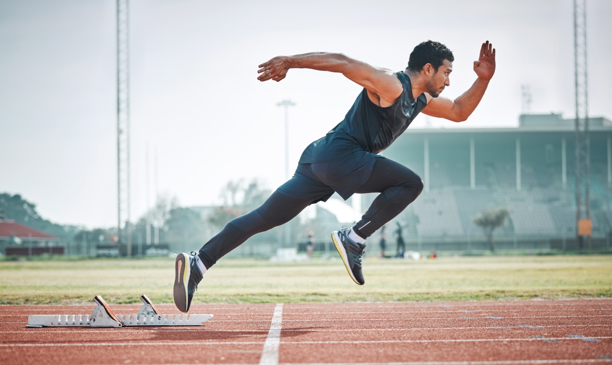 A sprinter in a black athletic outfit starts running on a track, captured mid-stride. The background features a stadium and clear sky.