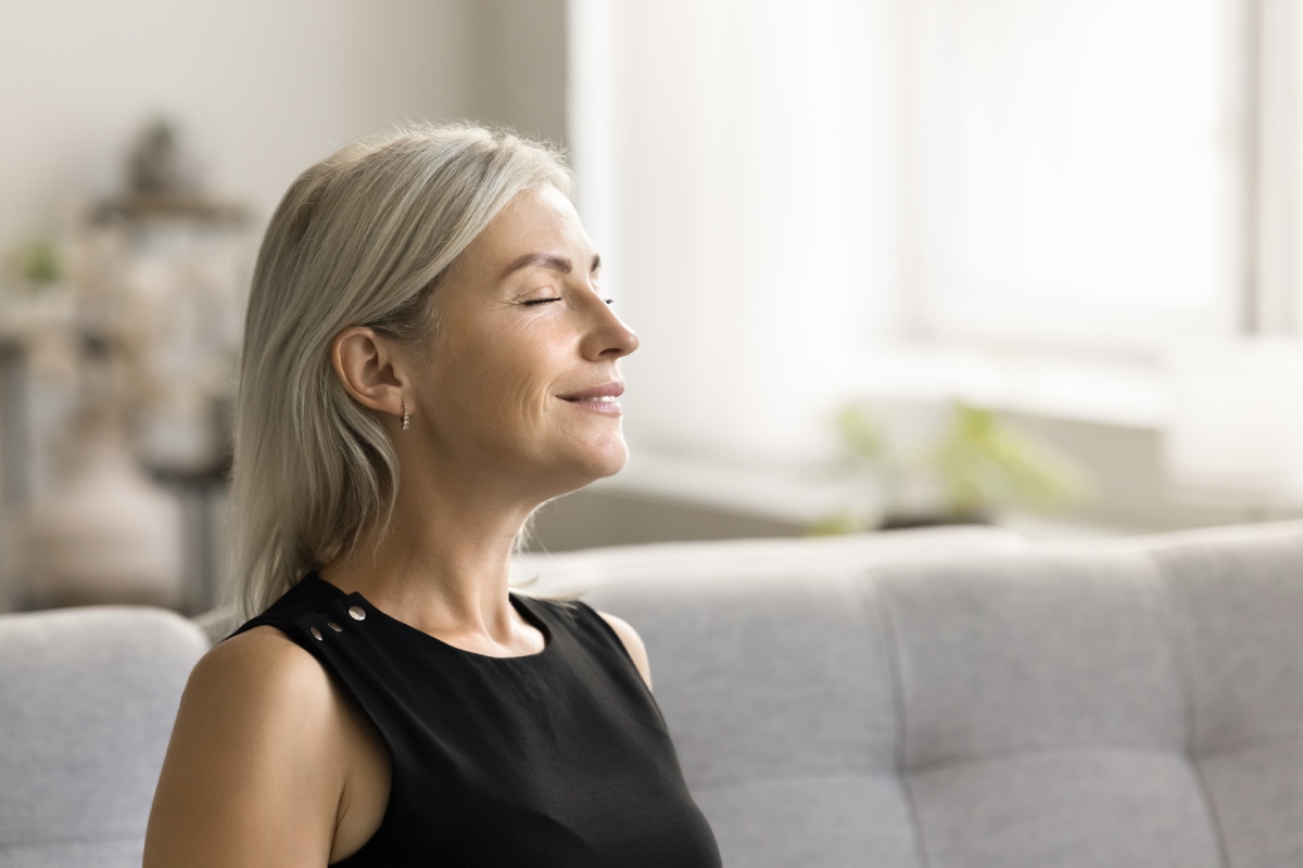 A woman with gray hair and a black sleeveless top sits on a couch with eyes closed, appearing relaxed in a well-lit room.