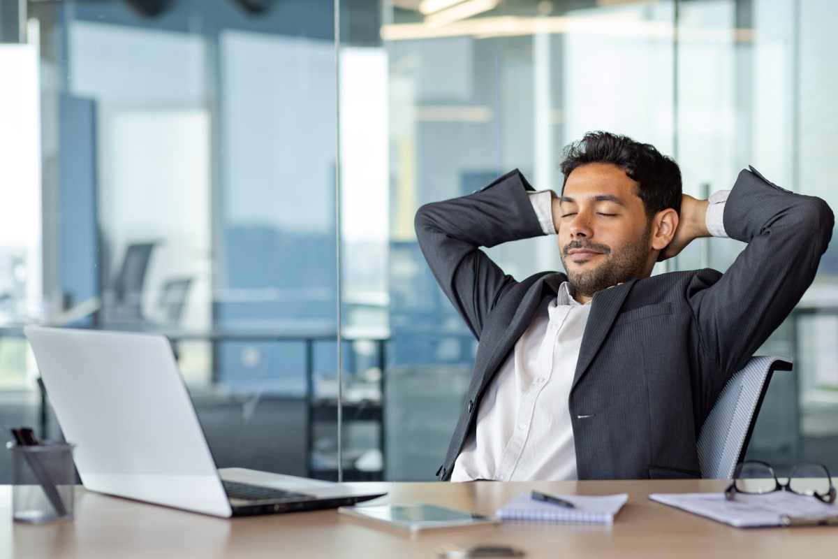 A man in a business suit sits at a desk with his hands behind his head, eyes closed. A laptop, phone, notebook, and pen are on the desk in front of him.