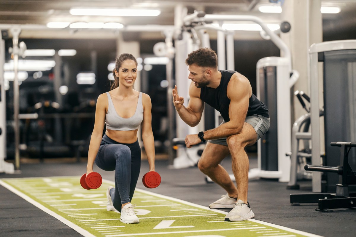 A woman performs a lunge exercise with red dumbbells in a gym while a man crouches beside her, giving instructions.