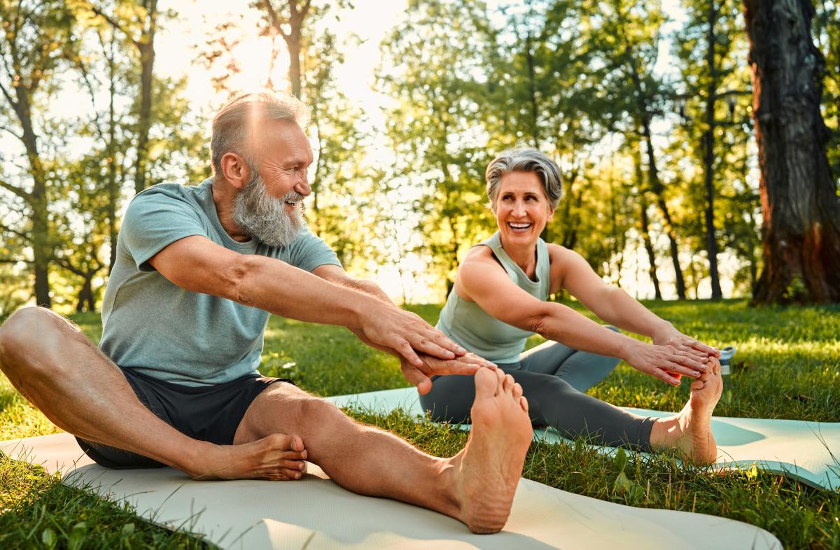 Two older adults, a man and a woman, performing seated forward bends on yoga mats in a sunny, wooded outdoor setting.