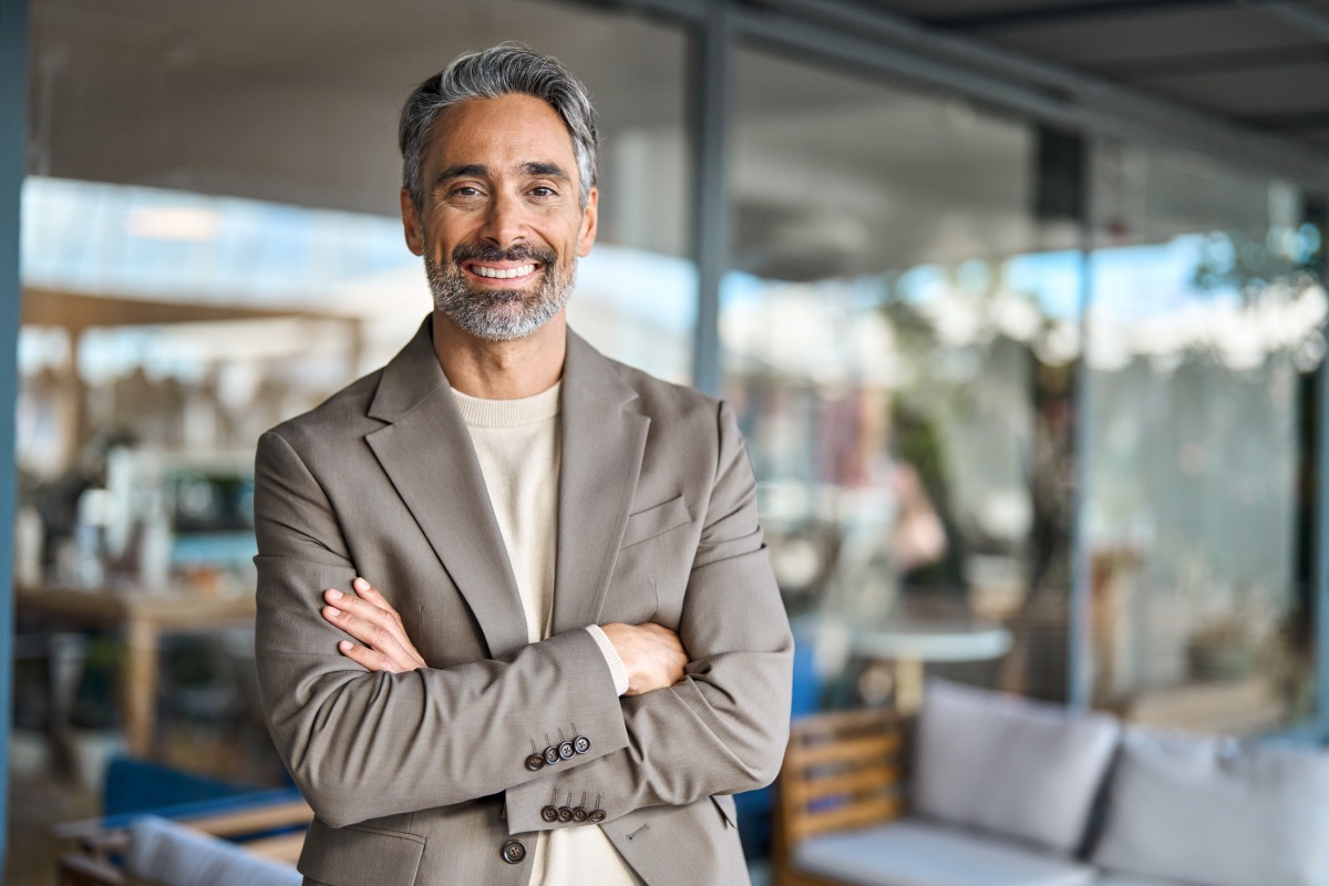 A smiling man with grey hair and beard stands with arms crossed in an outdoor seating area, wearing a beige blazer over a light shirt.