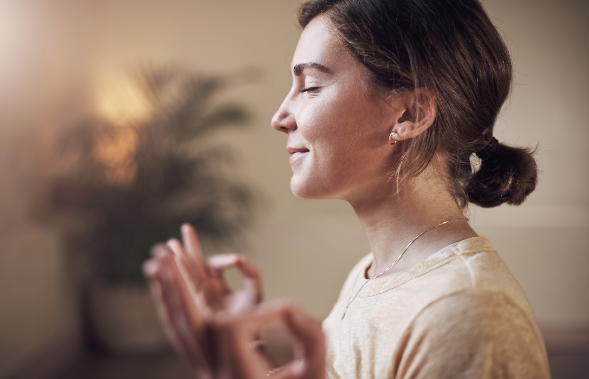 Woman with closed eyes sitting indoors, meditating, with her hands in a yoga mudra position. A blurred plant is visible in the background.