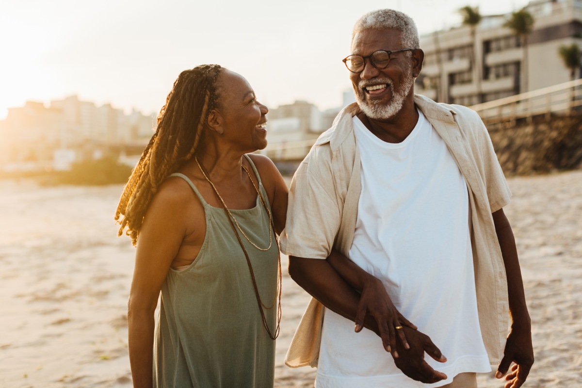 An elderly couple smiles and walks arm-in-arm on a beach near a city, with the sun setting in the background.