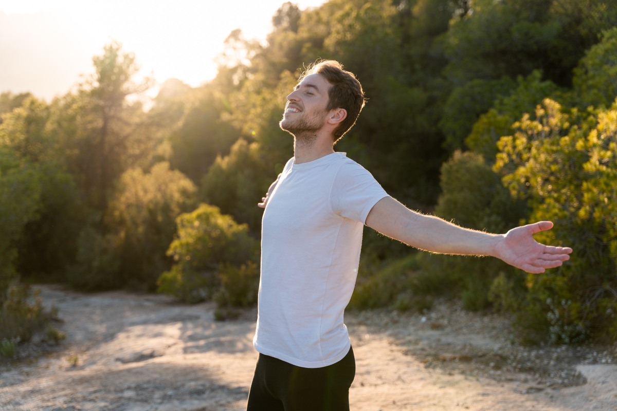 A man in a white t-shirt stands outdoors on a sunlit path, eyes closed, with arms outstretched, surrounded by trees.