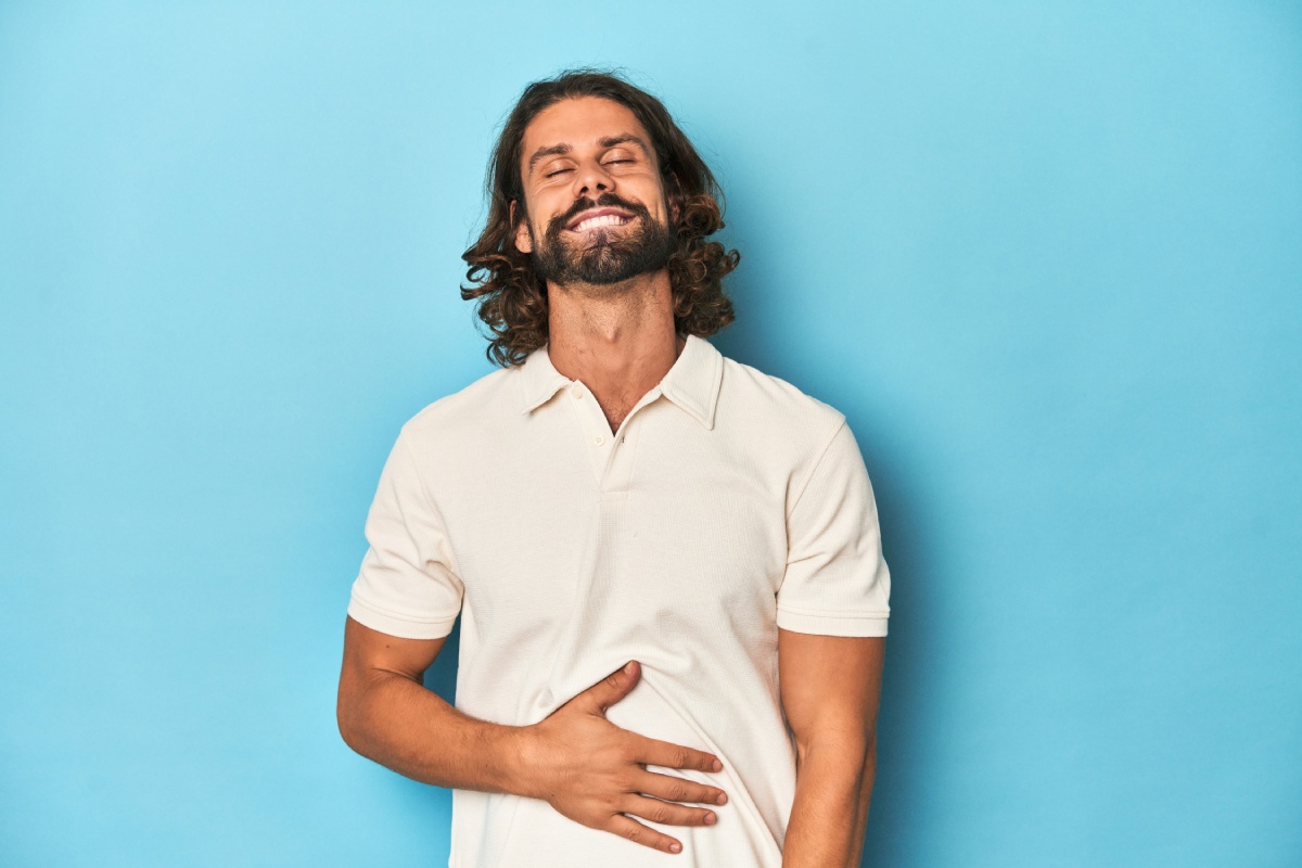 Long-haired man in a white polo, blue studio touches tummy, smiles gently, eating and satisfaction concept.
