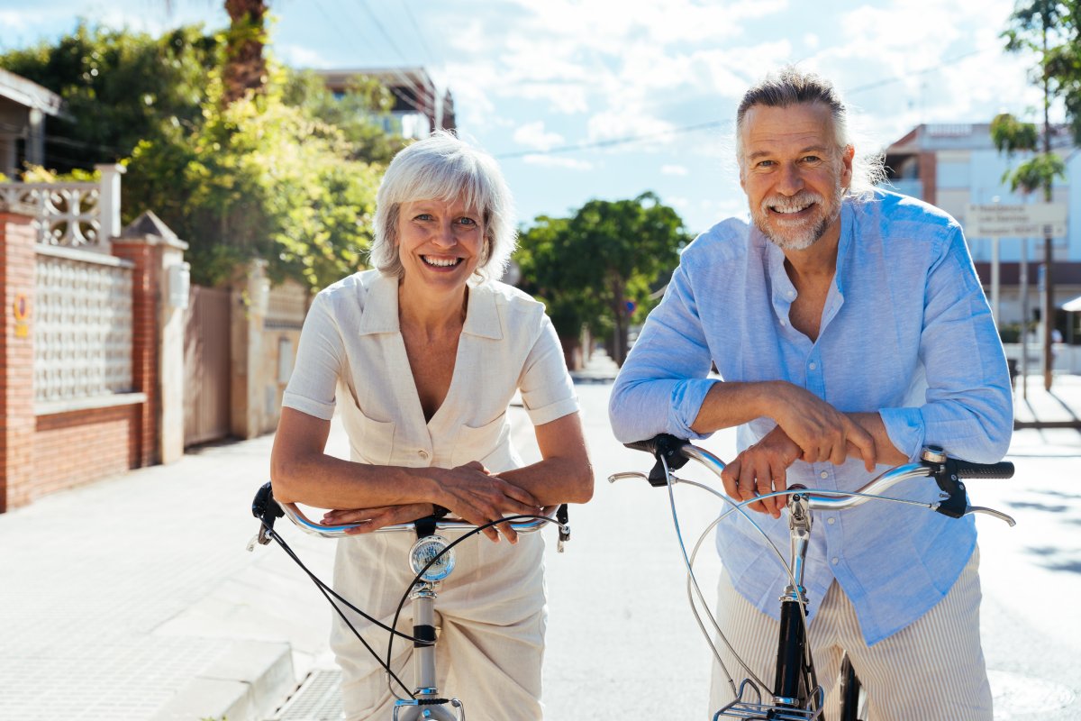 Two older adults, one woman and one man, stand outdoors with their bicycles on a sunny day, smiling and leaning on the handlebars. Trees and buildings are in the background.