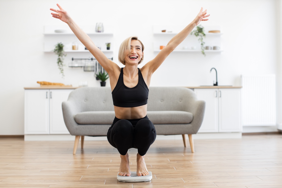 A woman in athletic wear is standing on a scale with arms raised, smiling in a bright living room. A grey sofa and kitchen area are in the background.