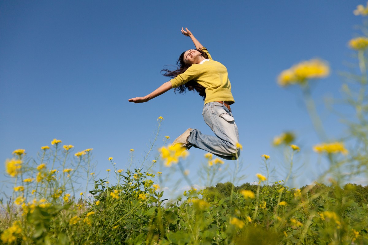 A person wearing a yellow shirt and jeans jumps joyfully in a field of yellow flowers under a clear blue sky.
