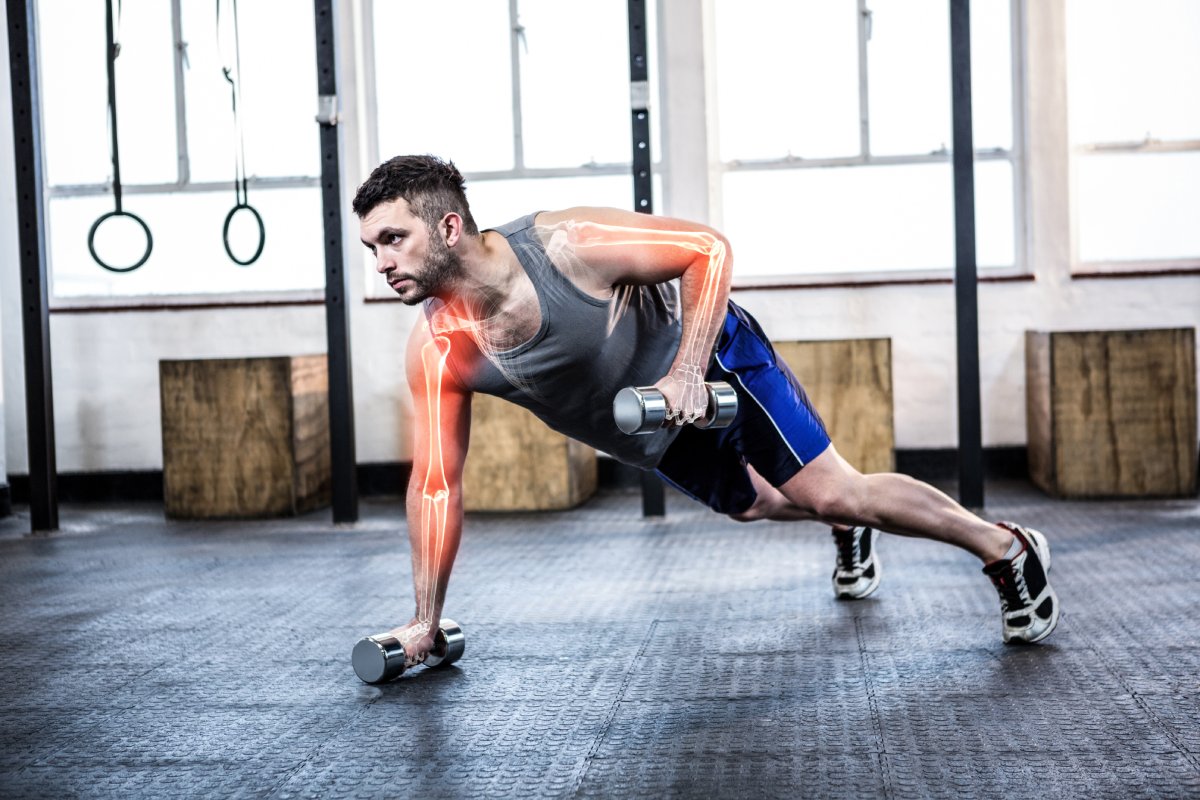 Man in a gym doing a one-arm dumbbell row with highlighted arm muscles, wearing a gray tank top and blue shorts.