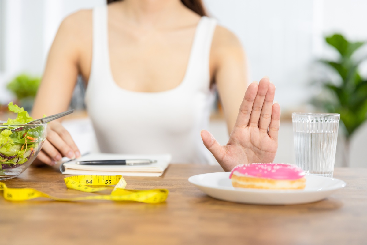 Person refusing a donut with a hand gesture, sitting near a salad, a glass of water, a notebook, and a yellow measuring tape.