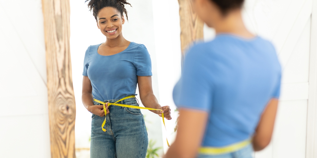 A person in a blue shirt and jeans uses a yellow measuring tape around their waist, smiling at their reflection in a mirror.