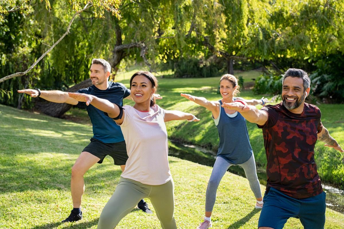 Four people practicing yoga outdoors in a park, standing with arms extended in a warrior pose, surrounded by trees and grass.