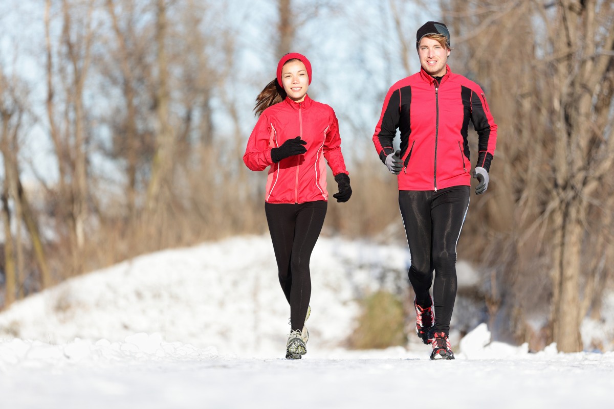 Two people jog on a snowy path wearing red jackets and black pants. Trees line the background.