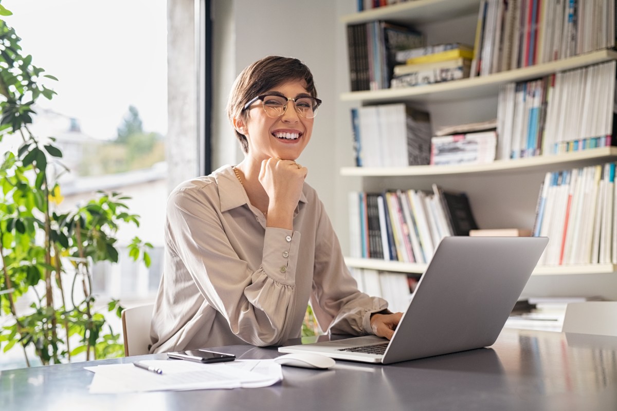 Person with glasses smiling at a desk, using a laptop, surrounded by books and papers, near a window with plants.