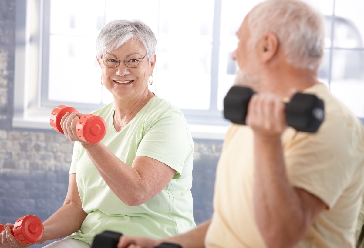 Two elderly individuals are exercising indoors with dumbbells. The woman wears a light green shirt and smiles, and the man, in a yellow shirt, is facing her.