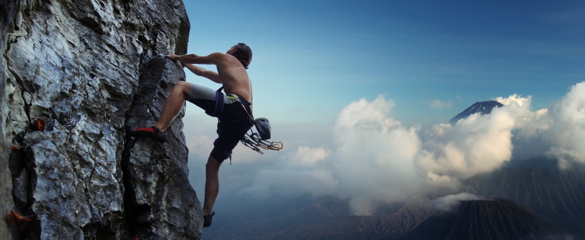 A man is rock climbing on a steep cliff with clouds and distant mountains in the background.