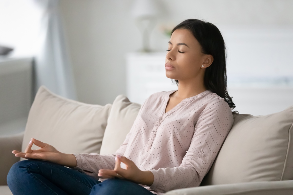 A person sits cross-legged on a couch with eyes closed and hands resting on knees, appearing calm and meditative.