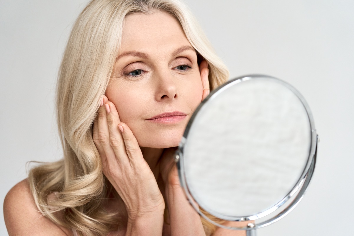 A woman with long blonde hair looks at herself in a round mirror, gently touching her face with her hands.