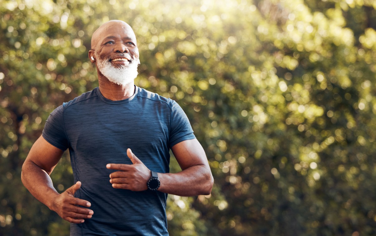 A man with a beard wearing a blue shirt and smartwatch is smiling while outdoors. Trees and sunlight are in the background.