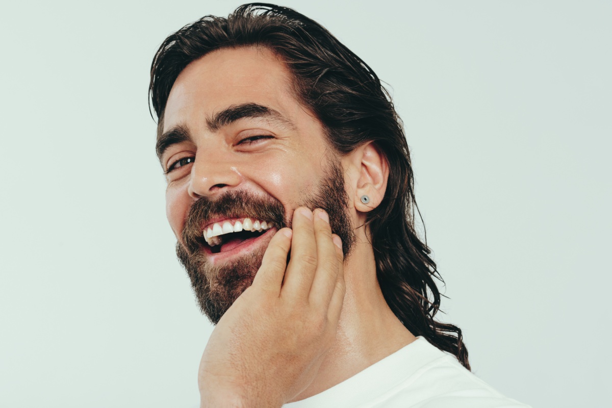 A man with a beard and long hair smiles brightly, touching his cheek with one hand.