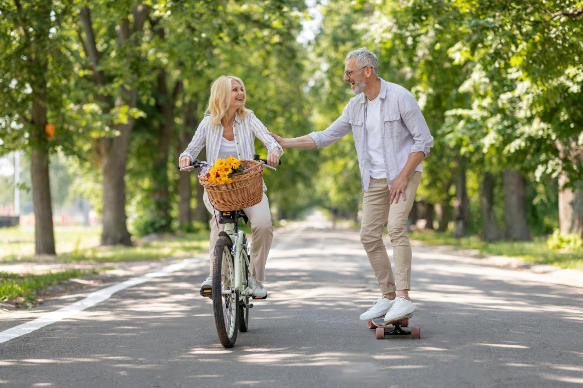 A man on a skateboard playfully interacts with a woman riding a bicycle with a basket of flowers on a tree-lined path.