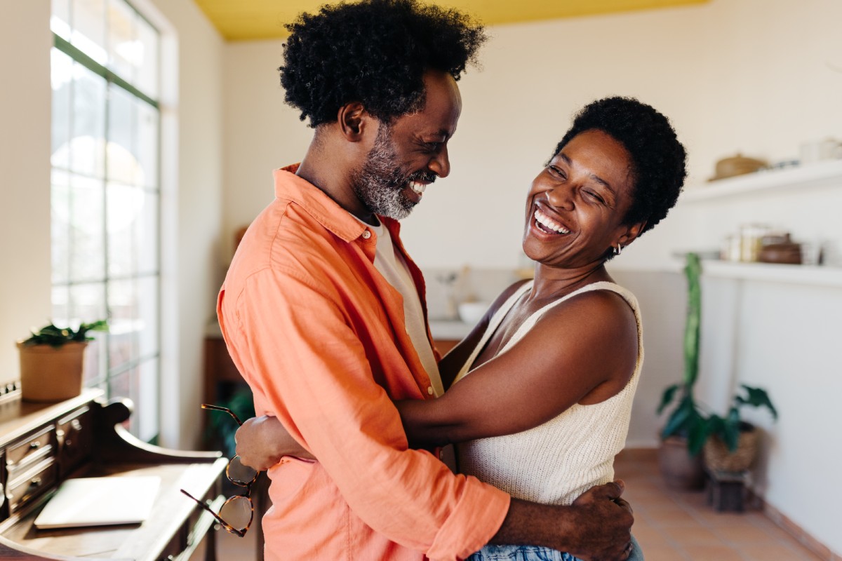A couple embracing and smiling at each other in a bright room with plants and a window in the background.