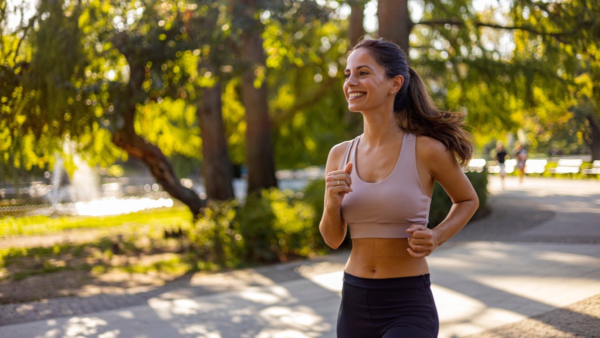 A woman jogs in a park on a sunny day, wearing a light sports top and dark leggings, with trees and a fountain in the background.