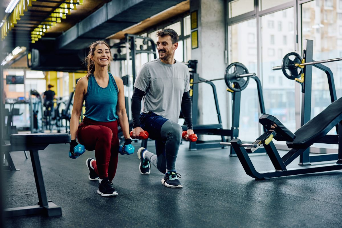 Two people perform lunges with dumbbells in a gym. They are wearing athletic clothing and appear focused. Fitness equipment is visible in the background.