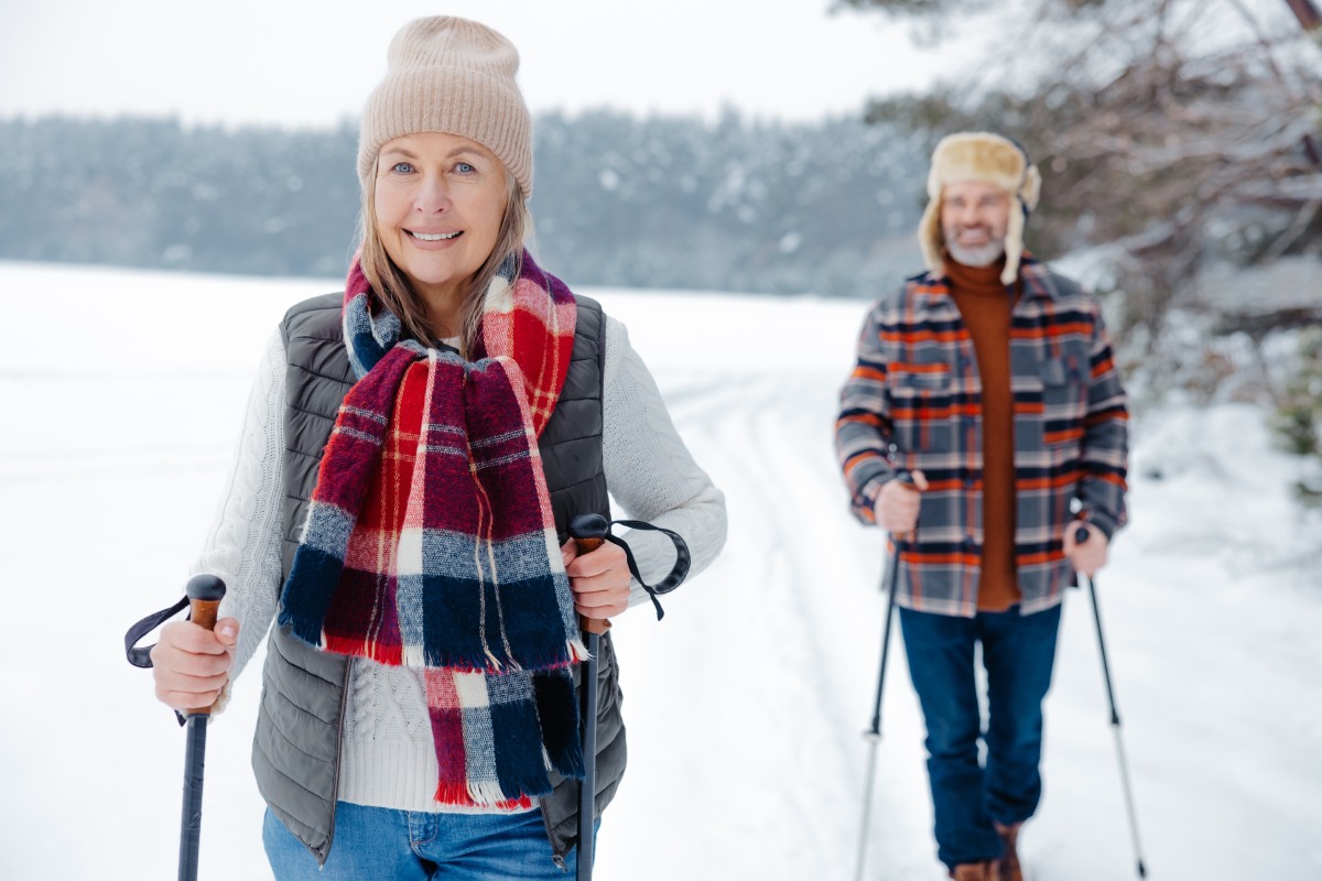 A woman and man are walking with ski poles on a snowy path. Both are wearing winter clothing, including hats and scarves. Trees and snow surround them.