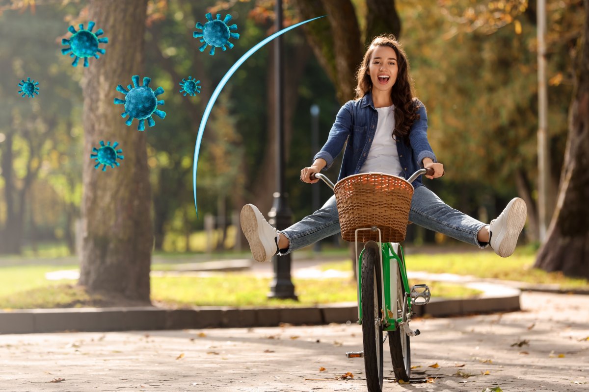 A smiling girl rides a bicycle with arms outstretched. Blue virus-like symbols are illustrated around her in a park setting.