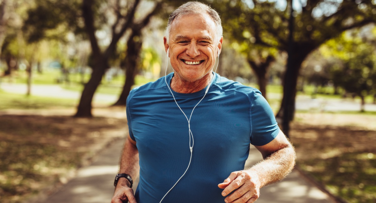 Older man jogging in a park, wearing a blue t-shirt and earphones, smiling, with trees and a path in the background.