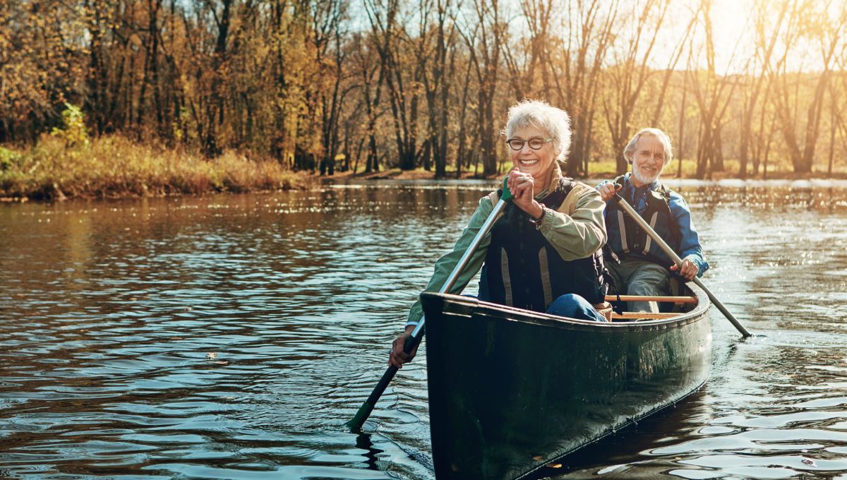 Two people paddle a canoe on a calm river, surrounded by trees and illuminated by sunlight.