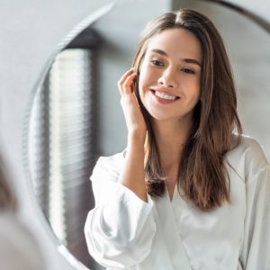 A woman in a white robe smiles while looking in a round mirror.