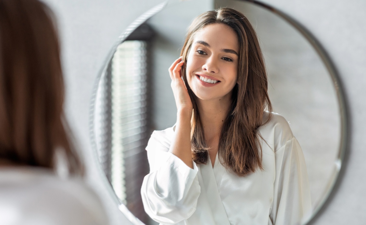 A woman in a white robe smiles while looking in a round mirror.