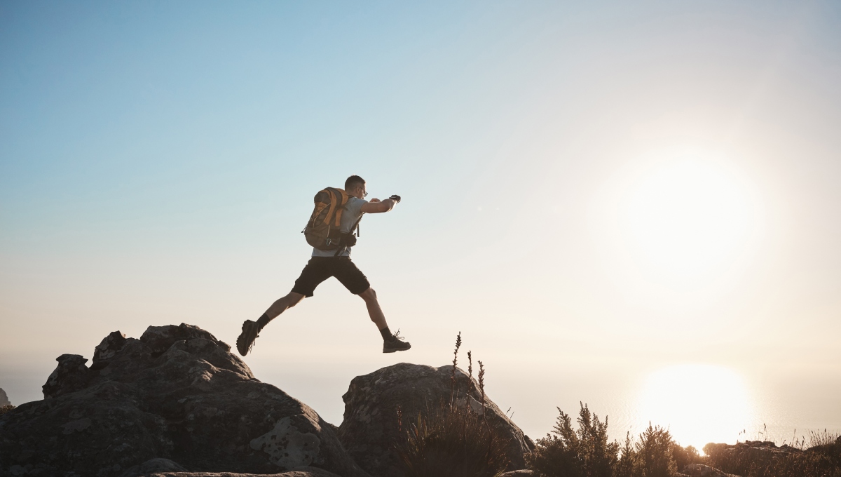 A person with a backpack jumps between rocks at sunset, with the sun low in the sky and a clear horizon in the background.