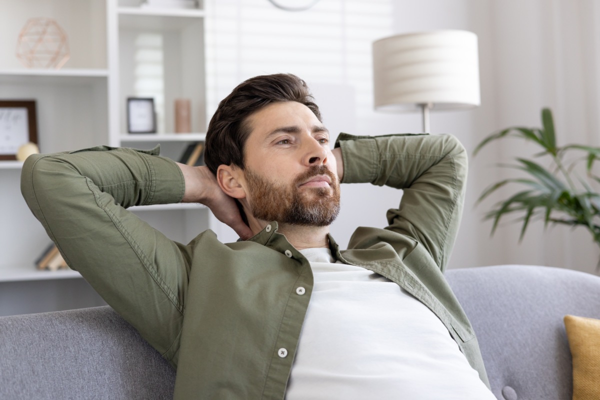 A bearded man in a green shirt relaxes on a gray couch with hands behind his head in a bright living room. A potted plant and lamp are in the background.