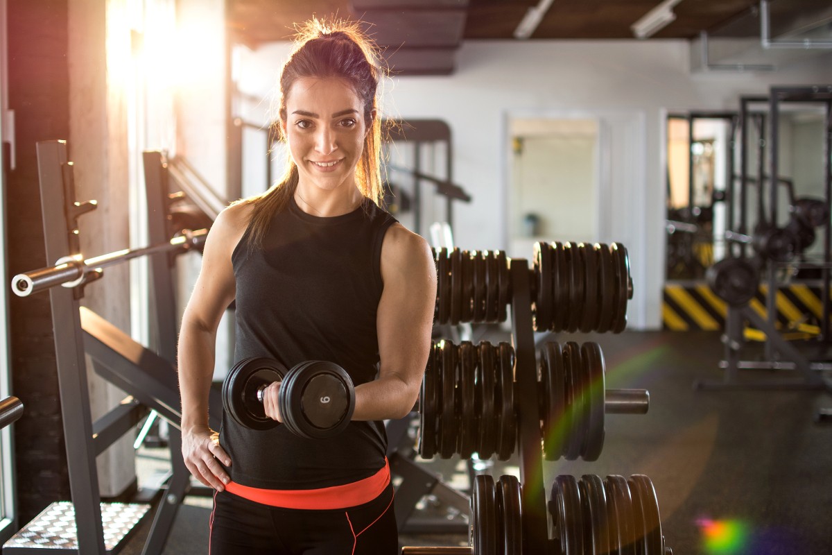 Person in a gym holding a dumbbell, standing near a rack of weights with sunlight streaming in.
